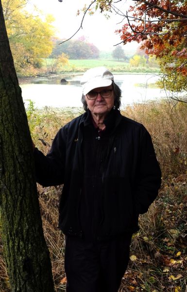 Portrait of the photographer, Richard Quinney. He is wearing a black jacket, eyeglasses, and a white baseball cap, and is standing with his right hand resting on the trunk of a tree. In the background is the pond surrounded by trees and plants turning fall colors.