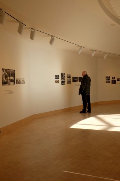 A man standing and observing photographs on display on the wall of the James Watrous Gallery. The photography is black and white, showing images of buildings, horses, and people, etc. Light from a window illuminates the floor behind the man.