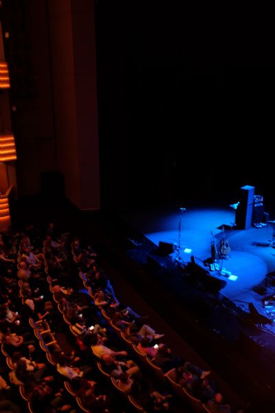 View from balcony looking down at the stage at the Overture Center. People are sitting in the ground floor seats. The stage is illuminated by blue spot lights, and amplifiers, microphones, and two guitars are on the stage. 