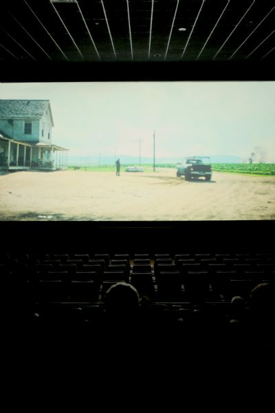View of a movie screen from the seats in the middle of a theater. There are a few people sitting in the seats in the foreground. On the screen is a scene of a building with a porch, a person and a truck on the dirt road in front. In the background is a field.