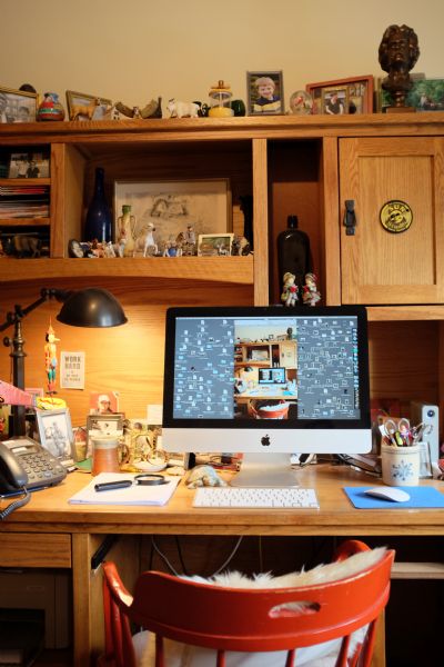View of a red chair in front of a desk with built-in shelves. In the center is a Macintosh computer and keyboard. A telephone, lamp, and magnifying lens are on the left. Photographs and figurines cover the desk space and shelves of the hutch above the computer. A small sign behind the computer reads: "Work hard and be nice to people." On the computer screen is an image of the same desk, computer, and chair.