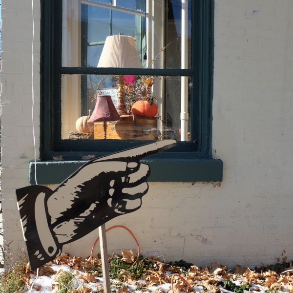 View of a sign in the shape of a hand in front of a window on a white brick wall. Leaves and snow are on the ground. Inside the window, two lamps, two chests, two pumpkins and decorative autumn foliage are lit by sunlight.