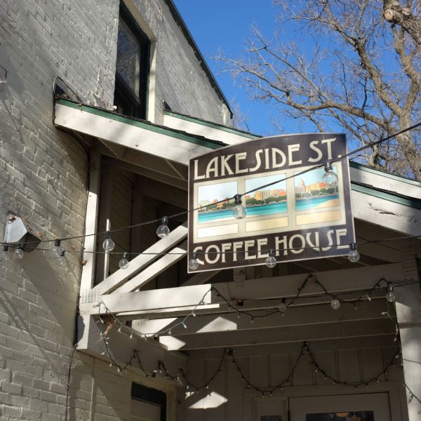 View looking up at decorative lights hanging from the beams of an entry roof. A sign on the side of the overhang reads: "Lakeside St. Coffee House." The sign also features a triptych of downtown Madison. 
