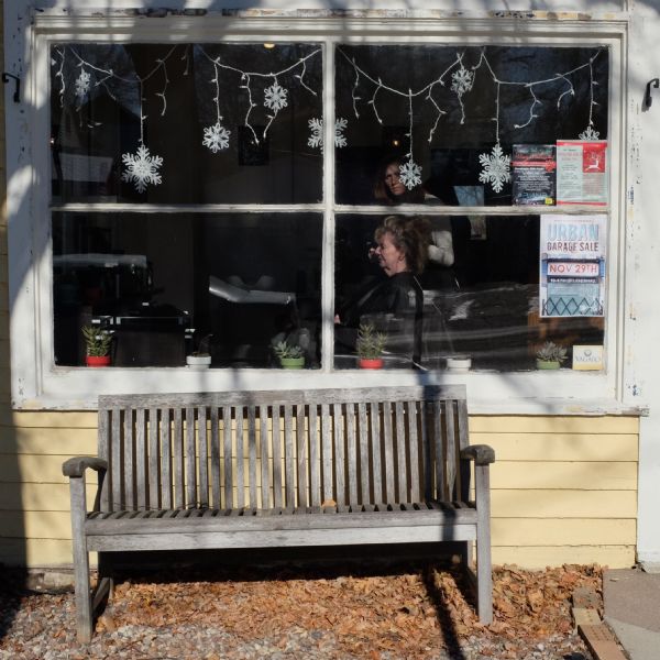 View of an old, wooden bench sitting in front of a large, front window of a beauty salon. Decorative snowflakes and lights are hanging on the inside of the window, along with three flyers, and small potted plants are along the windowsill. Inside, a woman is sitting in a chair as the hairdresser attends to her hair.