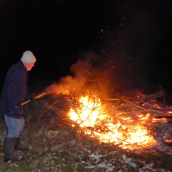 James Danky dressed in thick boots, a coat, gloves and hat, prods at a large fire with a rake. Several large twigs, sticks, and branches provide fuel for the fire. Smoke and embers rise into the air.