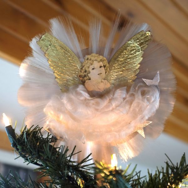 Close-up view of an angel tree topper on a Christmas tree. The angel is made of a paper cut-out with the image of a young girl with curly hair, shiny gold metallic wings, a fluffy, white flared dress, and a white plastic filament backing.