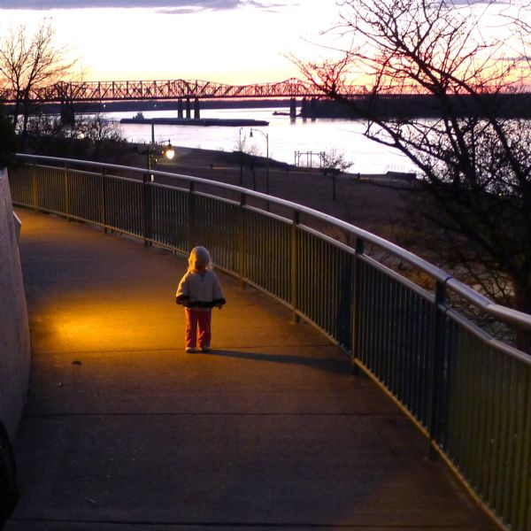 View of a child walking on a walkway with metal railings. There is a light shining down on her from a wall on the left. In the background is a slope going down to the the shoreline of a river. There is a bridge in the distance silhouetted against the sky.