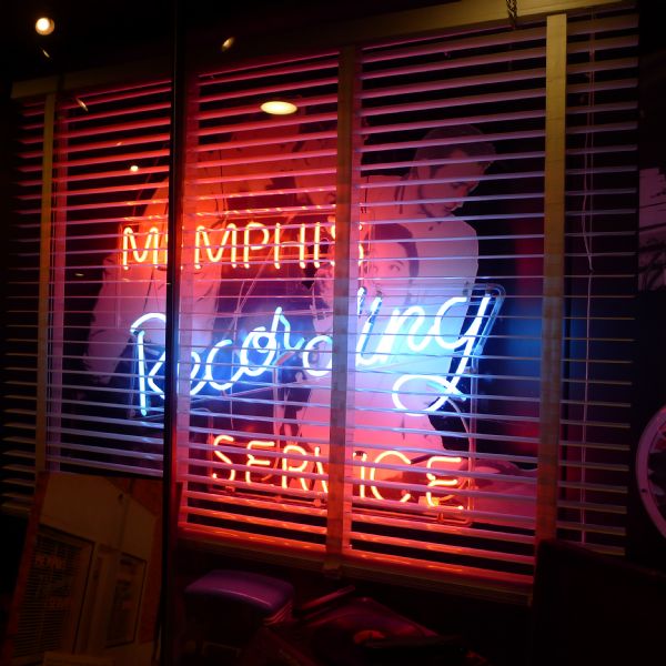A red and blue neon sign in a window with Venetian blinds reads: "Memphis Recording Service." Behind the window is a cardboard cutout of three men leaning over Elvis Presley, who is sitting.