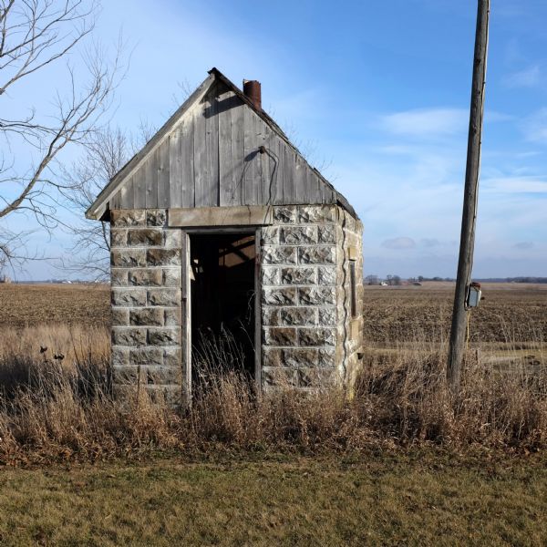 An abandoned stone and wood milkhouse sitting in tall grass in a field. Farmhouses and farm buildings are in the distance.
