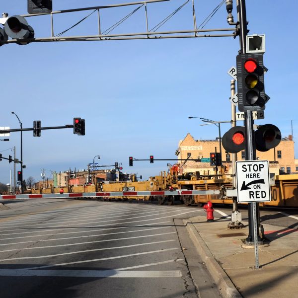 A cargo train is passing over Lincoln Highway. The railroad crossing gate is down and the traffic lights are red. Behind the train, old brick buildings line both sides of the street.