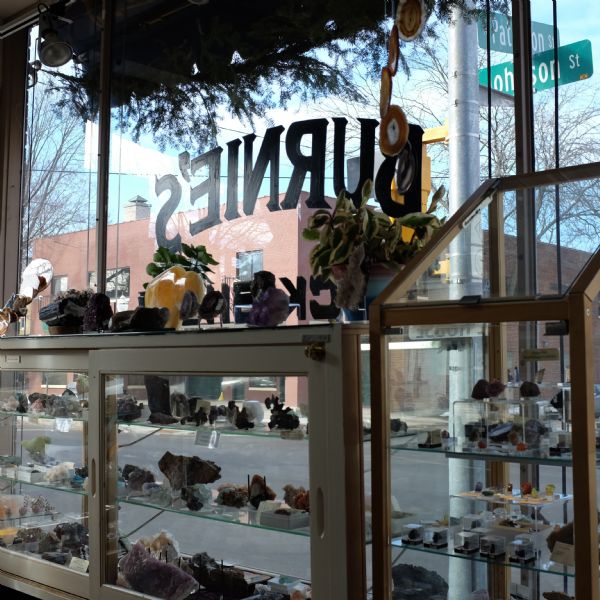 Inside view of display cases of rocks and geodes along the front windows of the store. "Burnie's Rock Shop" is painted in reverse on the window. Two street signs name the streets at the intersection of Johnson and Patterson streets. Across the intersection is a brick building.