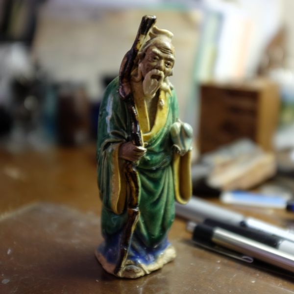 Close-up of a small, clay statue of Shou Lao, the Chinese deity of longevity, sitting on a desk near some pens.