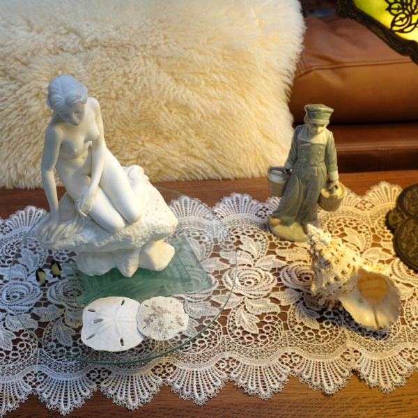 Several figurines, starfish, and shells sitting on top of a lace runner on an end table. The figurines include: a young boy carrying two pails of milk, two tiny statues of penguins, and a white figure of a naked woman, perhaps a copy of the Little Mermaid statue in Copenhagen.