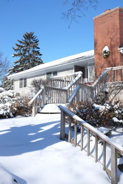 View up snow-covered stairs leading up to a porch of the photographer's house. A wreath is hanging on a large brick chimney near the front door. Bushes are on either side of the stairs, and trees are in the background.