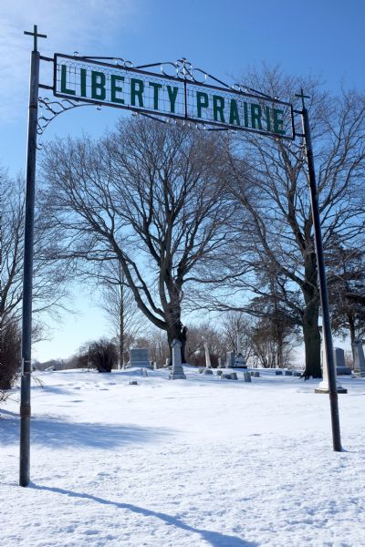 View of a metal sign to a cemetery that reads: "Liberty Prairie" on an ornate wire background. Grave markers in the snow are in the background.
