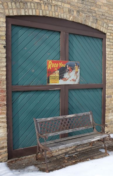 View of a wooden double door in the brick arch on the facade of a building. The door frame is painted brown and the four frames are painted teal. A worn, wooden bench surrounded by snow-covered ground is in front of the door. A painted metal sign, bolted into the door, keeps it closed. The sign features two women in 1950-style bathing suits water skiing, and reads: "Race You to Fluffy's, Wisconsin's Shangri-La! ... On the shores of Lake Ripley."