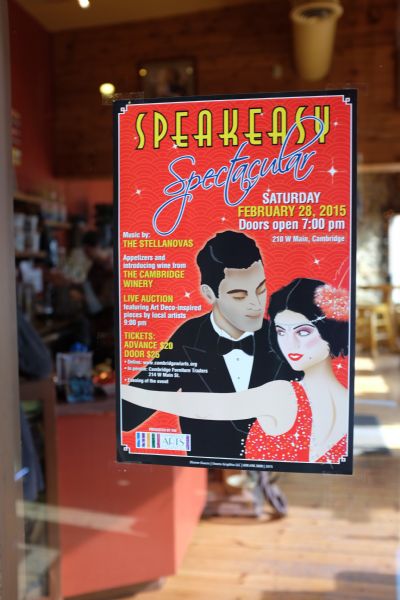 Close-up of a poster taped to the window of a cafe advertising an upcoming event, the "Speakeasy Spectacular." The poster features a man and woman in 1920s formal dance clothing. The interior of the cafe is in the background.