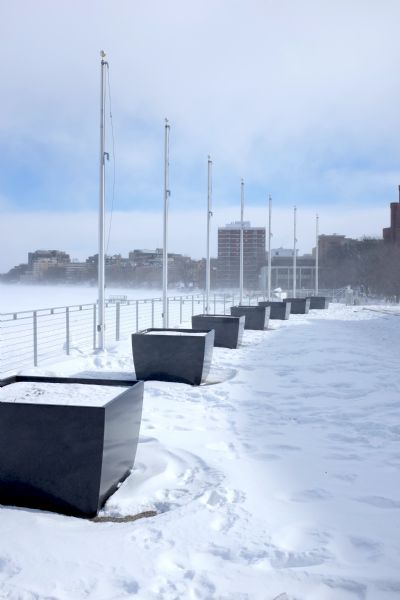 View of snow-covered Memorial Union Terrace, with large, dark-colored outdoor pots, and silver metal flagpoles spaced along the railing overlooking Lake Mendota. Snow is blowing across the lake towards buildings and trees along the shoreline in the background.