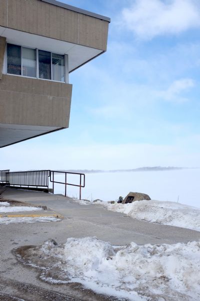 View of a corner of the Limnology Center on the shoreline of Lake Mendota. The building overhang is shading a walkway along the shore. A memorial stone and plaque for John "Vietnam" Nguyen sits to the left of the railing on the snowy shoreline, along with a bouquet of flowers. Snow blowing across the lake partially obscures Picnic Point in the far distance. 