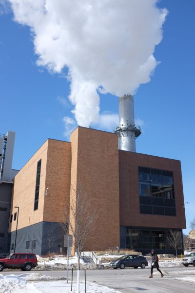 View of Charter Street Heating and Cooling Plant from North Mills Street. White smoke is billowing out of the stacks against a clear, blue sky. The streets are clear, but snow is covering the ground. A woman wearing a hooded coat is walking along the sidewalk.