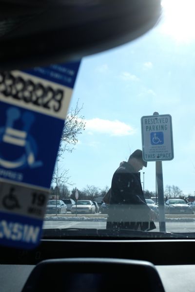 View looking out through a car windshield towards a parking lot. The dashboard, part of the rear view mirror, and a handicap parking sticker are inside the car. Outside is a man wearing glasses, a hat, and coat walking in front of the car, just behind a Reserved Parking sign.
