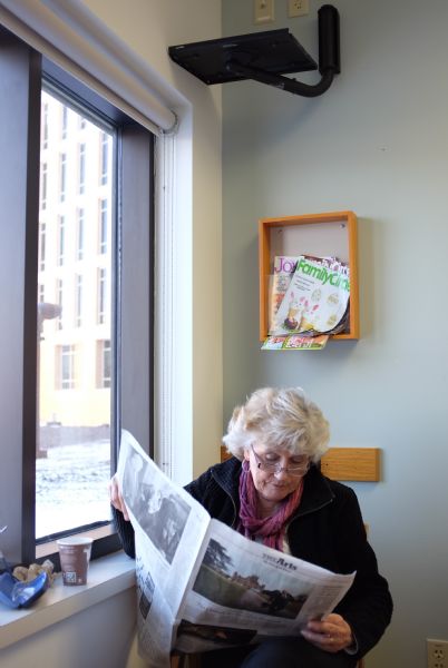 Solveig, the photographer's wife, sitting in a chair by a window reading a newspaper. A coffee cup is sitting on the window sill next to her, and magazines are in a display rack above her head.