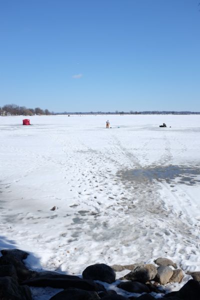 View from rocks shoreline of people walking or fishing on the snow- and ice-covered lake near a red ice fishing hut. Other fishing huts are in the distance. Trees and homes are along the opposite shore. 