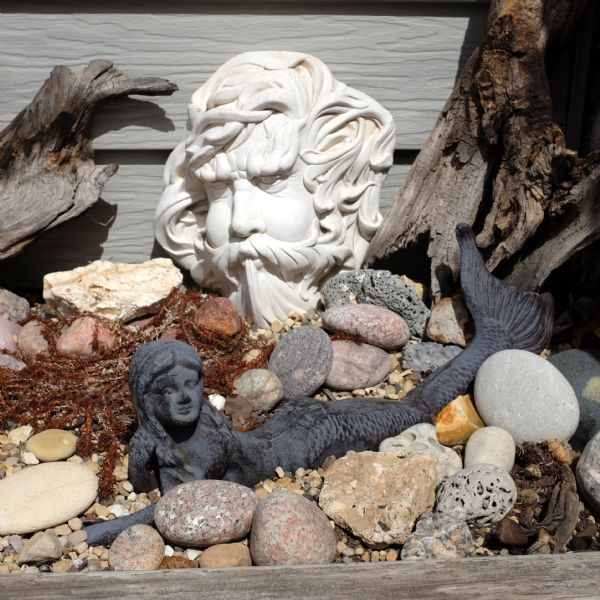 Two miniature statues are sitting in a rock garden set against a wall. One of the statues is of white stone with the face of a man with a mustache blowing in the wind. The other statue is of black stone in the shape of a mermaid lying on her side. Rocks, gravel, and two pieces of driftwood surround the statues. 