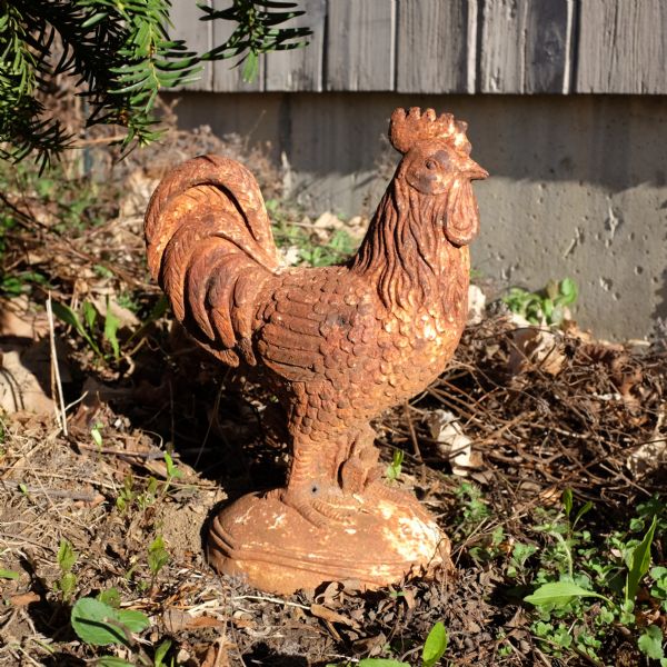 A red statue of a rooster sitting in the dirt in front of the foundation of a house.