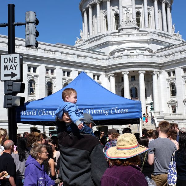 View of a crowd of people on the square, with the Wisconsin State Capitol in the background. One man is carrying a young boy on his shoulders. A tent over a stand at the market has a sign that reads: "Winning Cheese." 
