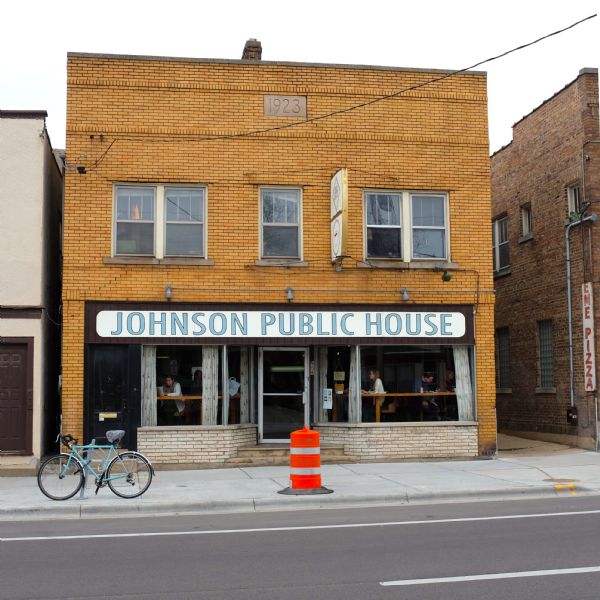 View across street towards a bicycle and traffic barrel standing on the sidewalk in front of a coffee shop. A sign above the first floor windows reads: "Johnson Public House." A date sign embedded into the building above the second floor windows reads: "1923." Women and men are sitting on chairs at high counters set inside the window.