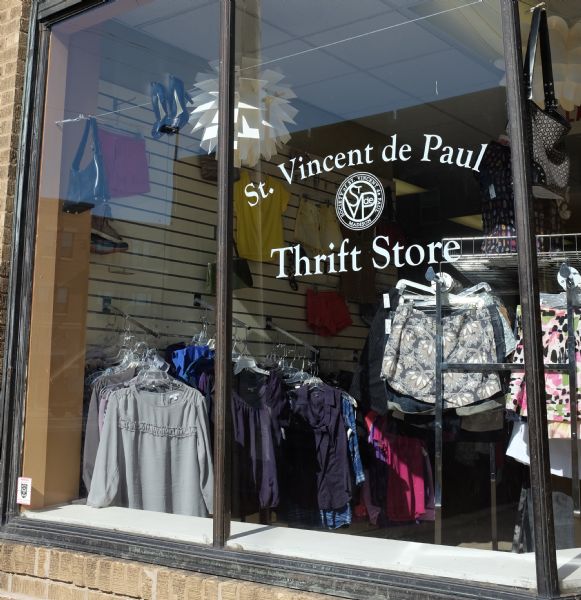 Exterior view through a window of the inside of St. Vincent de Paul Thrift Store. Shorts, purses, women's tops, and a pair of shoes are on display.