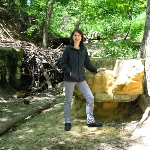 View of a young woman standing with her left hand resting on a rock outcropping, and her right hand in her jacket pocket. Trees are growing on the hill rising up behind her. A pile of branches is wedged between rock formations on the left.