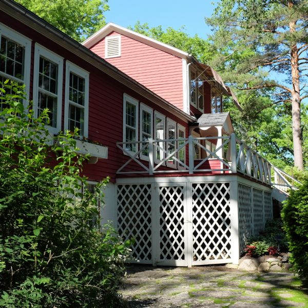 Exterior view of a red and white house surrounded by decorative bushes and trees. Stairs on the right are leading up to the porch and the main entrance. There are window boxes with blooming flowers. Awnings provide shade for the windows on the top floor. 