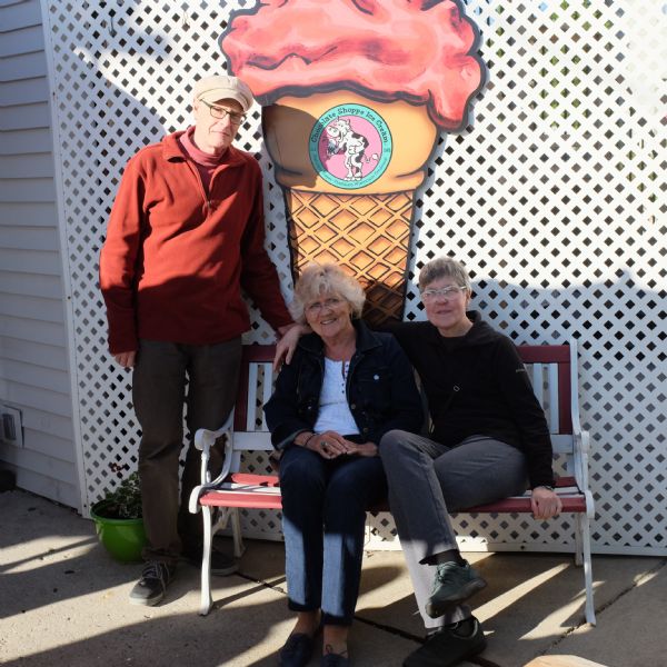 Group portrait of two women and a man (Paul Buhle) posing outdoors. The two women are sitting on a bench, and the man is standing next to them on the left. Behind them is a white lattice wall with a sign that reads: "Chocolate Shoppe Ice Cream."