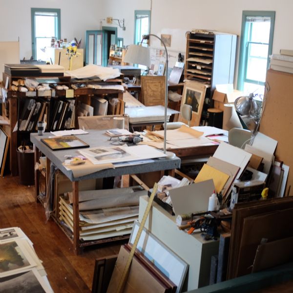 Slightly elevated view of a studio, with etchings, blank canvases, rolls of paper, and various tools covering every surface of the numerous tables and shelves in the studio. Three windows provide light.
