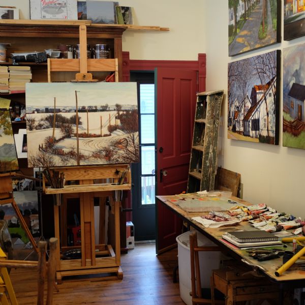 Larry Welo's paintings and etchings hanging on the wall in his studio gallery. One of the paintings is of a snow-covered landscape, and is displayed on a painter's easel in front of a bookcase filled with books, and painting and etching tools. A work table along the right wall has tubes of paint, tools, and notebooks, and behind the table a ladder splattered with paint is leaning against a wall next to a door which is slightly ajar.