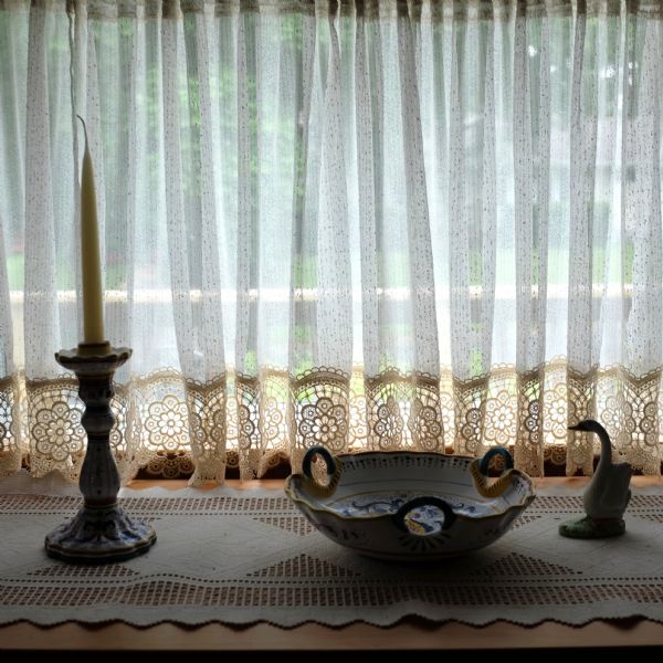 Interior view of sheer curtains edged in lace covering a window. On the lace runner covering the ledge along the window are decorative items, including a candle in a candlestick holder, a bowl, and a swan-shaped vase.
