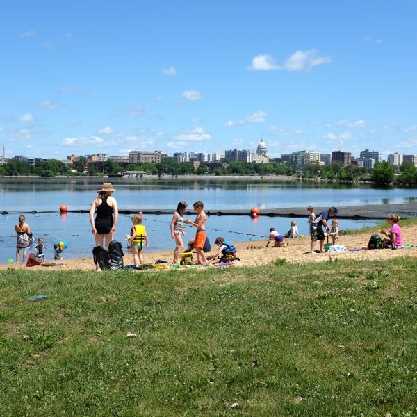 Children and adults at a small, sandy beach on the bay. Two woman are watching over the children as they play in the water or on the beach. The skyline of downtown Madison, including the the Wisconsin State Capitol, is in the distance.