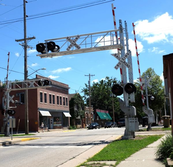 View down sidewalk towards a railroad crossing. Storefronts are along the sidewalk across the street, with cars parked along the curb in front.