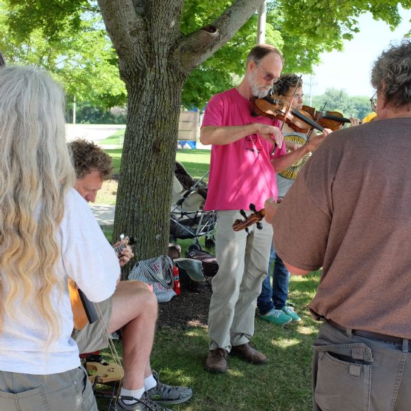 A group of men and women are standing and sitting under a tree playing string instruments.