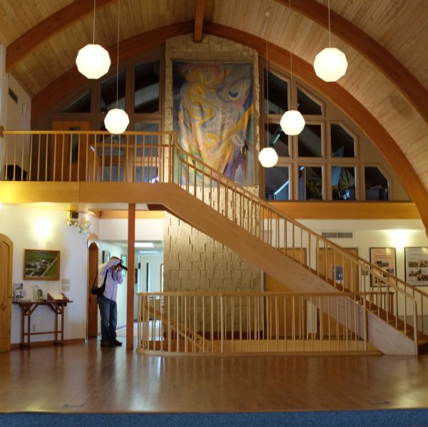 Indoor view of the Michael Fields Agricultural Institute. The room is arched, and has large windows on the second floor balcony, with a cloth painting hanging on the stone wall between the large windows. A wooden stairway goes up to the second floor, and another set of stairs goes down to the basement. A man is standing in front of the stairs to the basement taking a photograph with a camera.