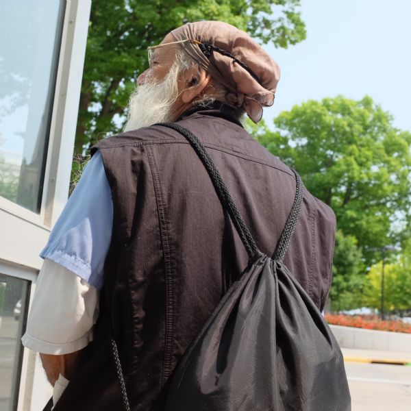 View of the back of an elderly man with a full white beard. He is wearing a backpack, eyeglasses, a cloth bandana on his head, two short-sleeved shirts, and a brown vest.