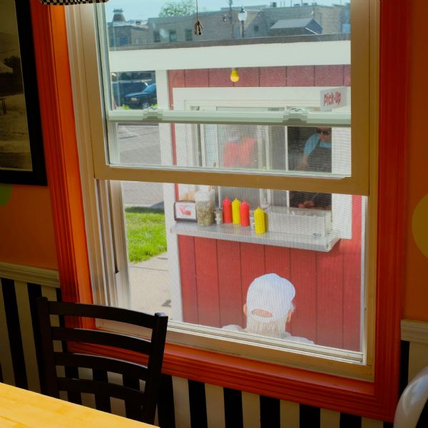 View from inside looking out through a window. In the foreground is a table and a chair in front of the window, and the view through the window is of a man waiting in front of a hamburger stand. Two people are working on a grill inside the stand, and condiments and napkins are on the metal counter in front of the pick-up window.