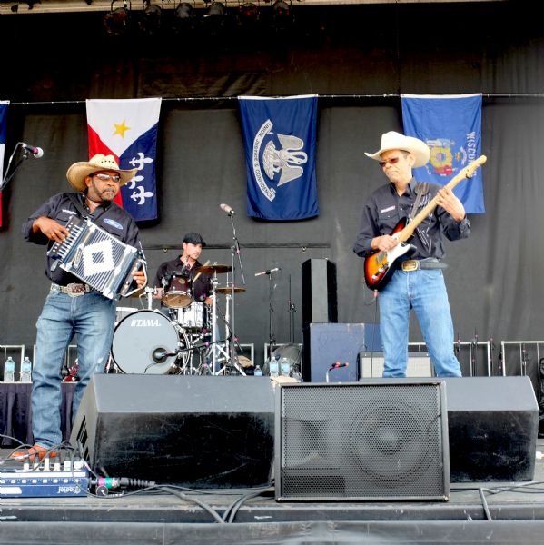View from audience towards a band playing on a stage. The men are playing an accordion, guitar, and drums. The man on the accordion is Jeffery Broussard. Behind the group are hanging four flags, from France, Acadiana, Louisiana and Wisconsin.