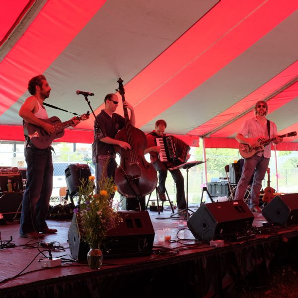 Four men are standing on a stage and playing instruments under a red and gray striped tent. Flowers are sitting in a vase at the front of the stage.