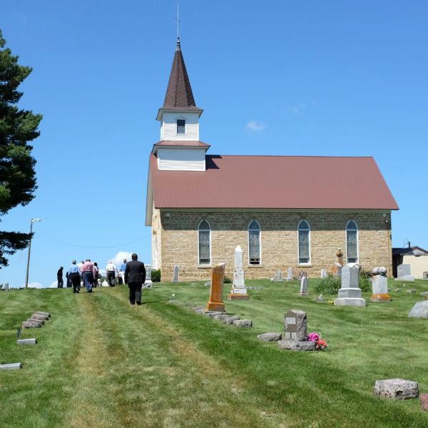 View up slope towards men and women walking along a grass path through a cemetery towards a church. The church is a one-room brick building with a steeple. 