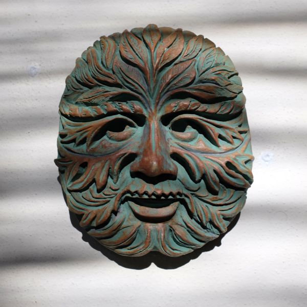 Ceramic mask of the Green Man hanging on a white stucco wall.