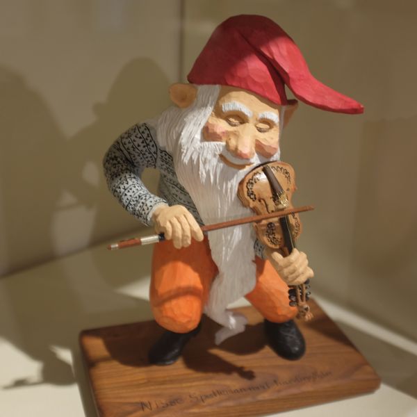 Carved wooden figure of a Nisse playing a fiddle. The figure has a long white beard and is wearing a red cap. Norwegian words carved into the base of the figure read: "Nisse Spelleman med hardingfele."