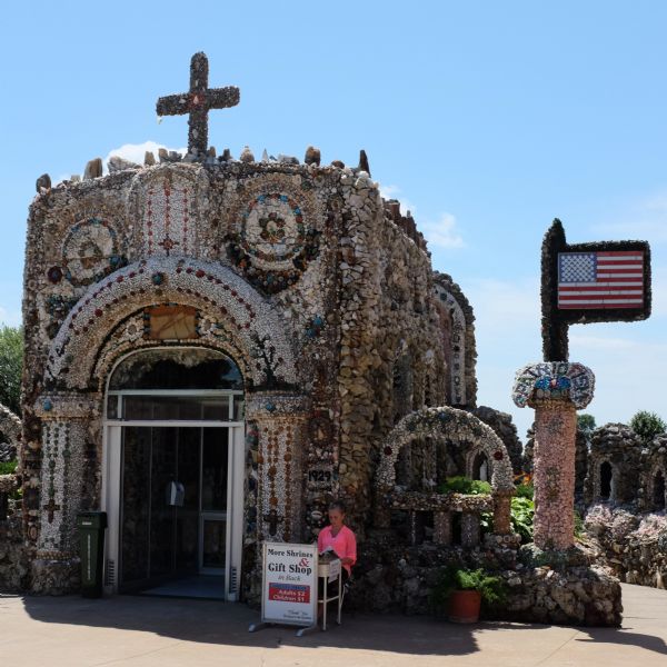 A small shrine made out of stone, mortar, colorful rocks and found objects. The modern door is made of glass and metal. A large stone cross is at the top of the shrine. A flagpole made in the same style as the building has an American flag, and the word "Patriotism" is set in mosaic just below. A young woman next to the entrance is sitting and reading a book behind a sign advertising the shrine, gift shop, and suggested entrance fee to the Grotto.